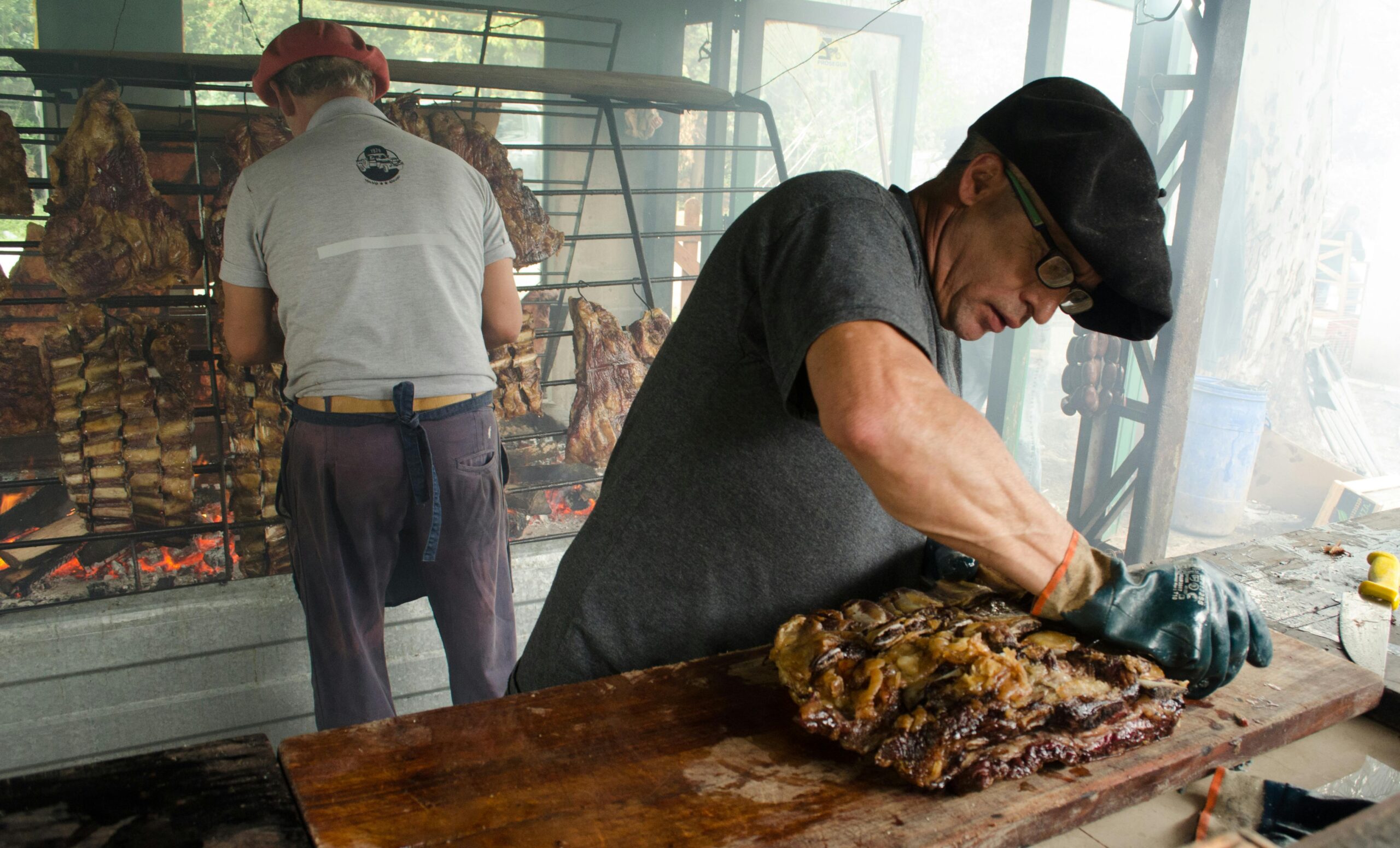 Two men preparing meat for barbecue on an outdoor grill with smoke rising.